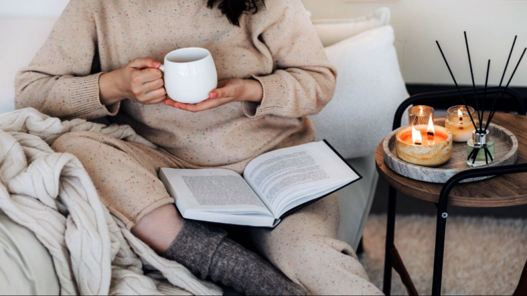 Cozy self-care moment with a person holding a warm mug of tea while reading a book on a couch, surrounded by soft blankets and lit candles.