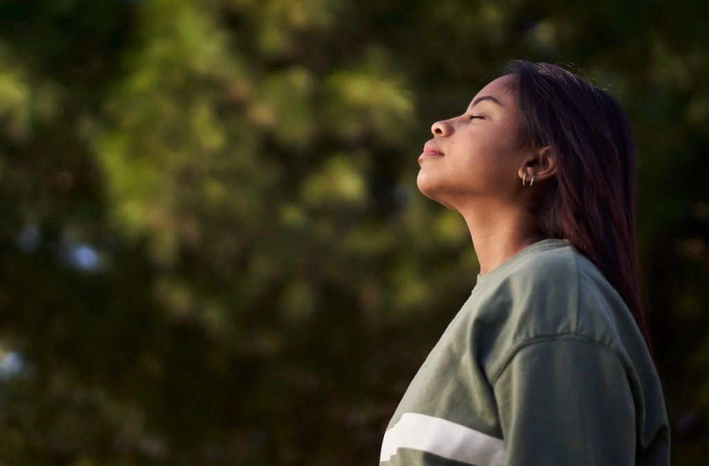Woman standing outdoors with eyes closed, taking a deep breath in a peaceful natural setting surrounded by greenery.
