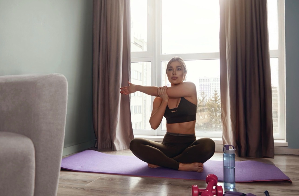 Woman stretches in a dimly lit room with cozy couch and dumbbells, water